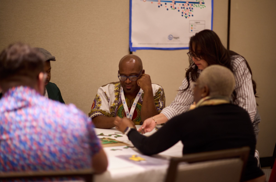 Five people sitting around a circular table that has papers spread across the top. They have contemplative looks on their face and one person is leaning across the table. Behind them, a large white piece of paper is taped to the wall.