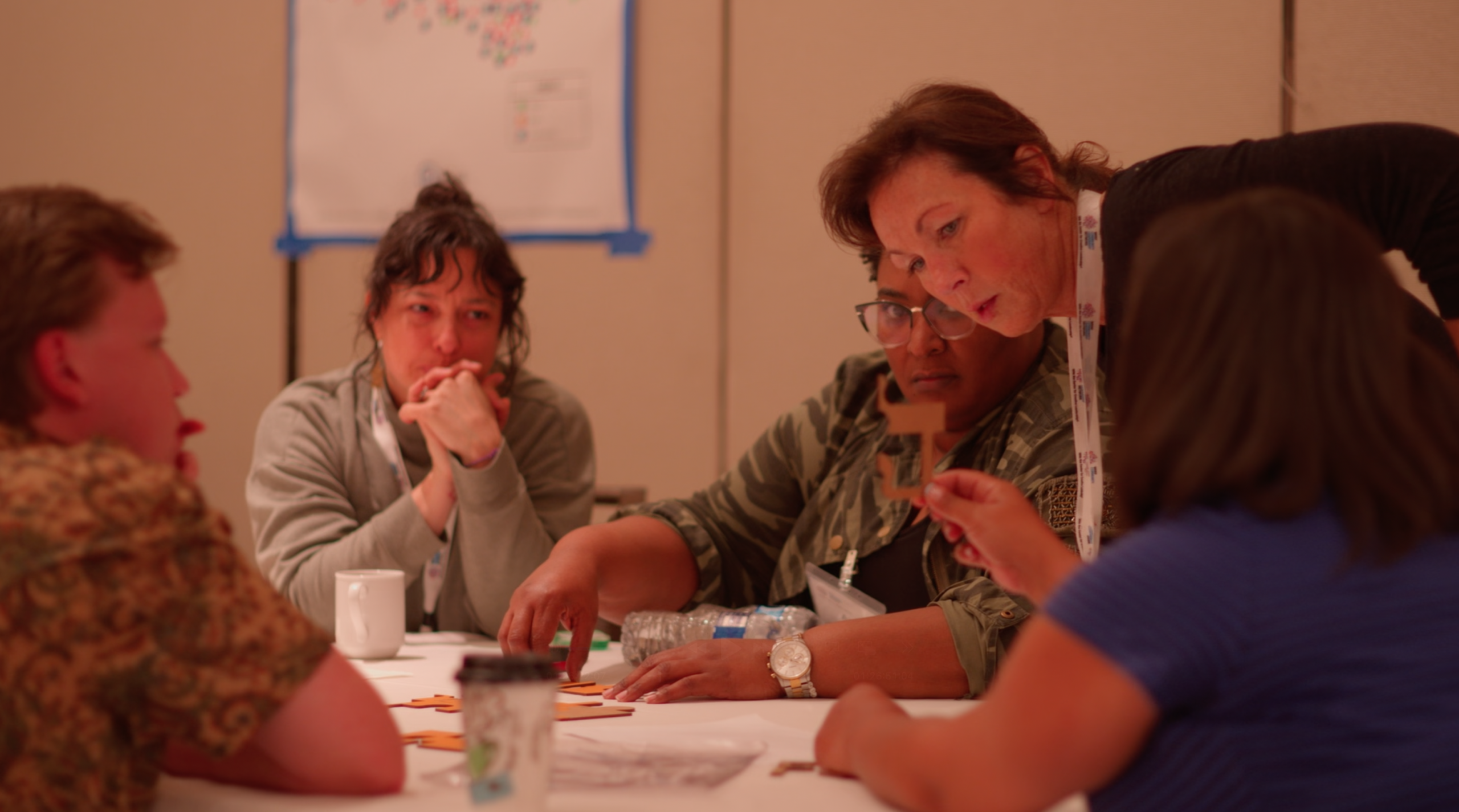 Five people sitting around a table that  has papers spread across the top. They have contemplative looks on their face and one person is leaning across the table.
